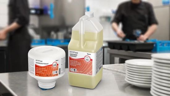 Keystone machine dishwaser liquid and solid dishwasher detergent sat on worktop in a dishroom with plates stacked up on the right of the image