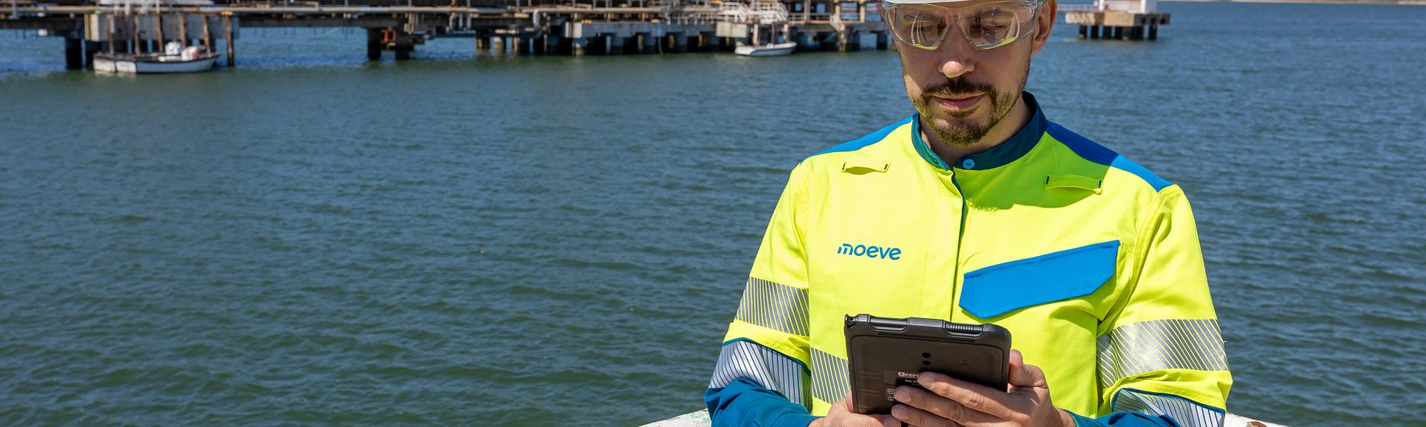 Industrial worker in high‑visibility safety gear using a tablet on a platform near the water, with an offshore facility in the background.
